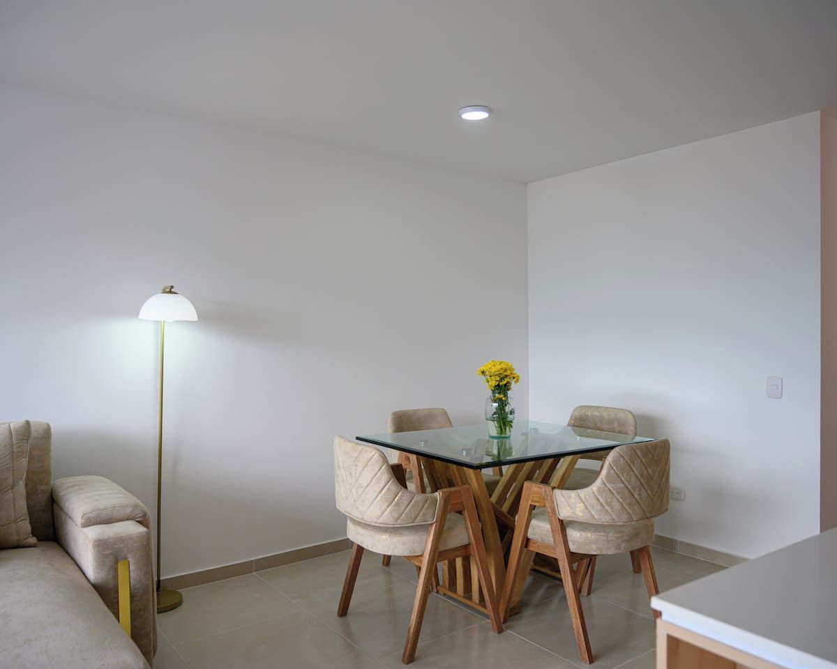 Ein Essbereich mit einem Glastisch, Holzstühlen, einer Stehlampe und einer Vase mit gelben Blumen.