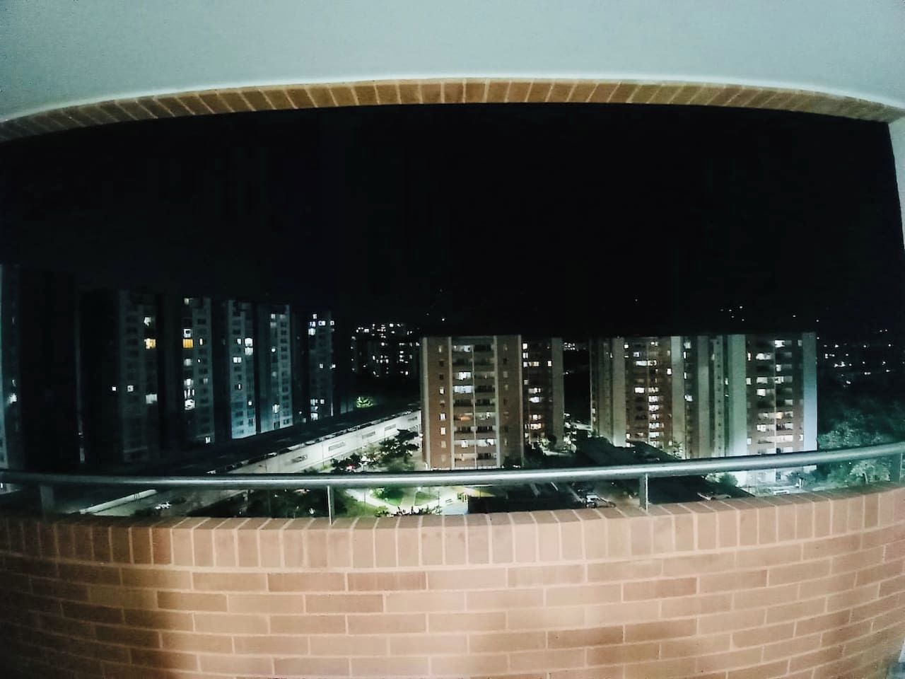 A nighttime view from a balcony with a brick wall, overlooking a cityscape with tall buildings and a road.