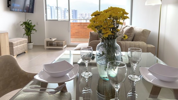 Un salon moderne avec une table à manger en verre pour quatre personnes, un vase rempli de fleurs jaunes et une vue sur la ville à travers de grandes fenêtres.