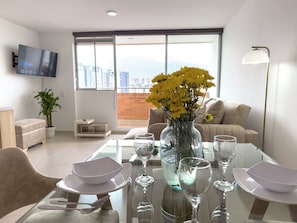 Un salon moderne avec une table à manger en verre pour quatre personnes, un vase rempli de fleurs jaunes et une vue sur la ville à travers de grandes fenêtres.