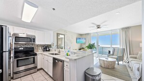 A modern kitchen with stainless steel appliances, a dishwasher, and a large island with a sink. There is a dining area with a round table and chairs, and a balcony with a view. - Beach Club Condos  (Gulf Shores)