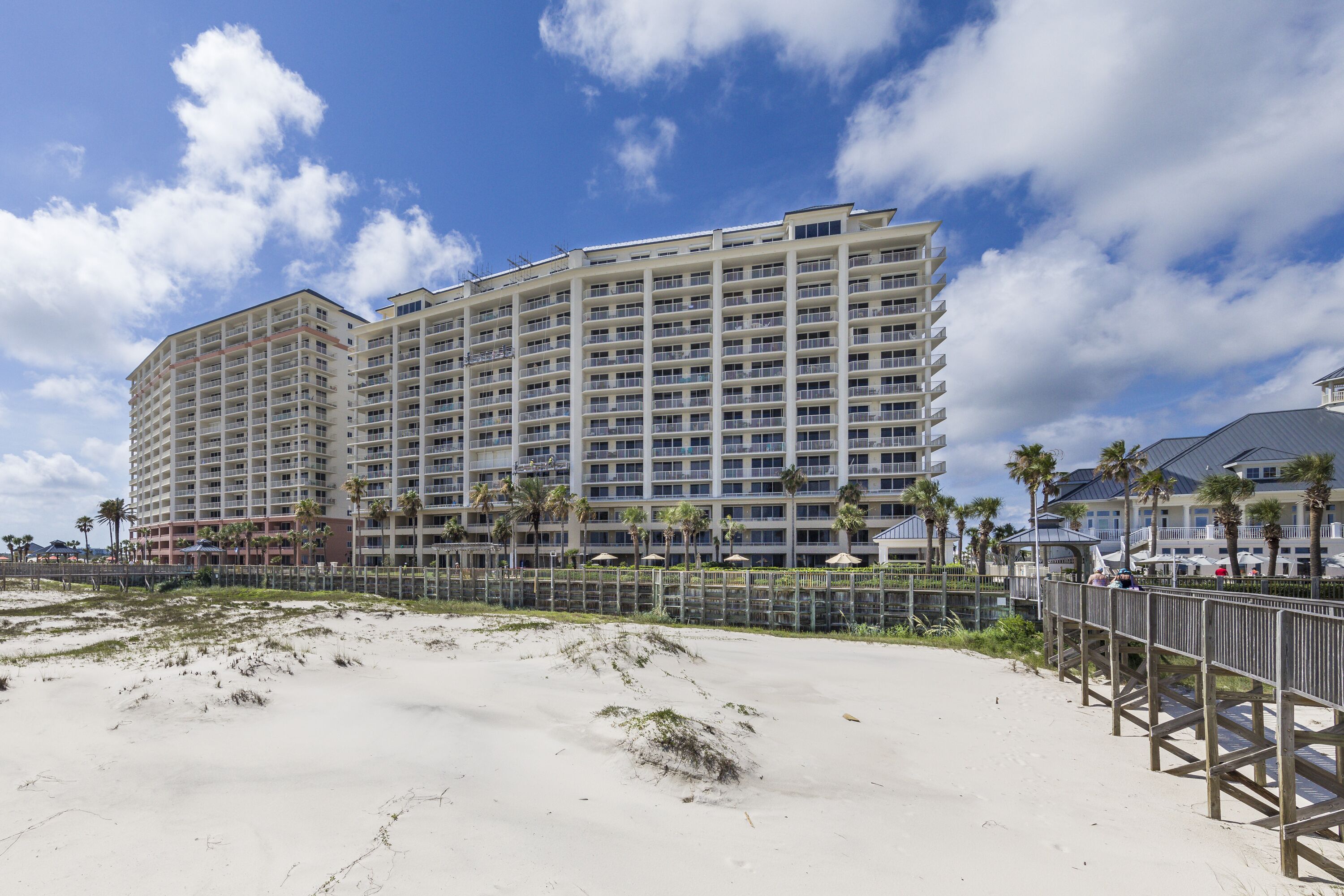 Un hotel grande con varios pisos y balcones, ubicado cerca de una playa arenosa con un paseo marítimo de madera.