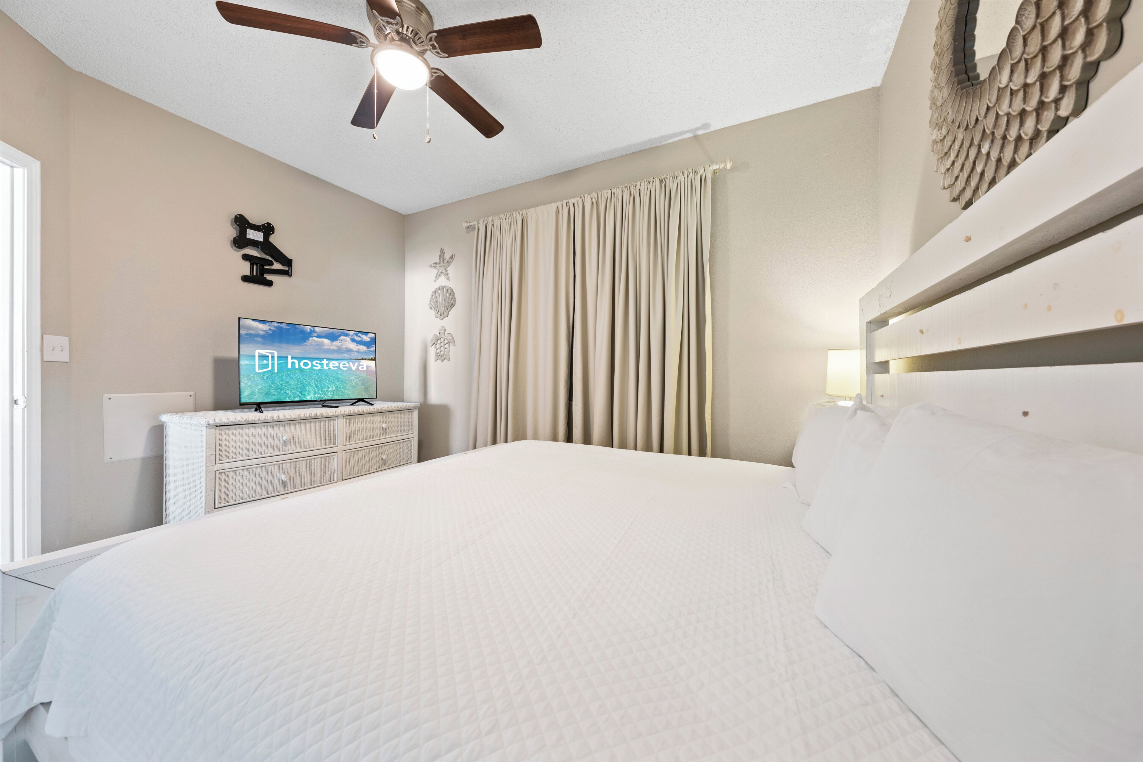 Ein Schlafzimmer mit einem Bett, einem Deckenventilator, einem Fernseher auf einer Kommode und einem an der Wand befestigten Regal.
