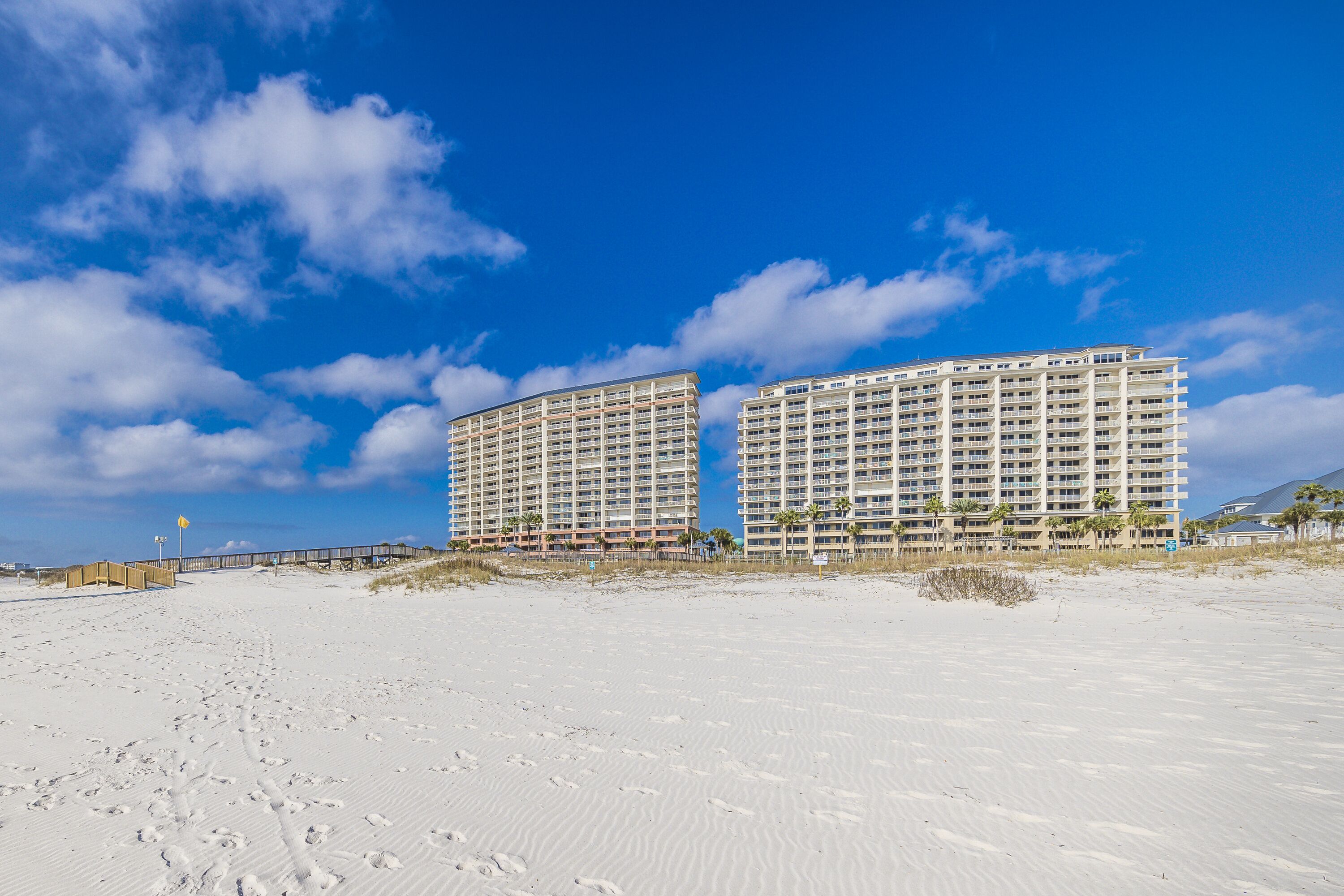 Un hotel frente a la playa con varios pisos y balcones, una playa de arena en primer plano y un cielo azul despejado.