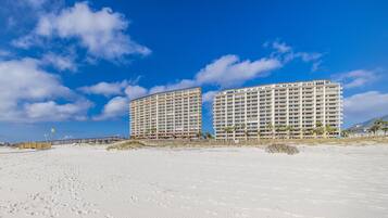 A beachfront hotel with multiple floors and balconies, a sandy beach in the foreground, and a clear blue sky.