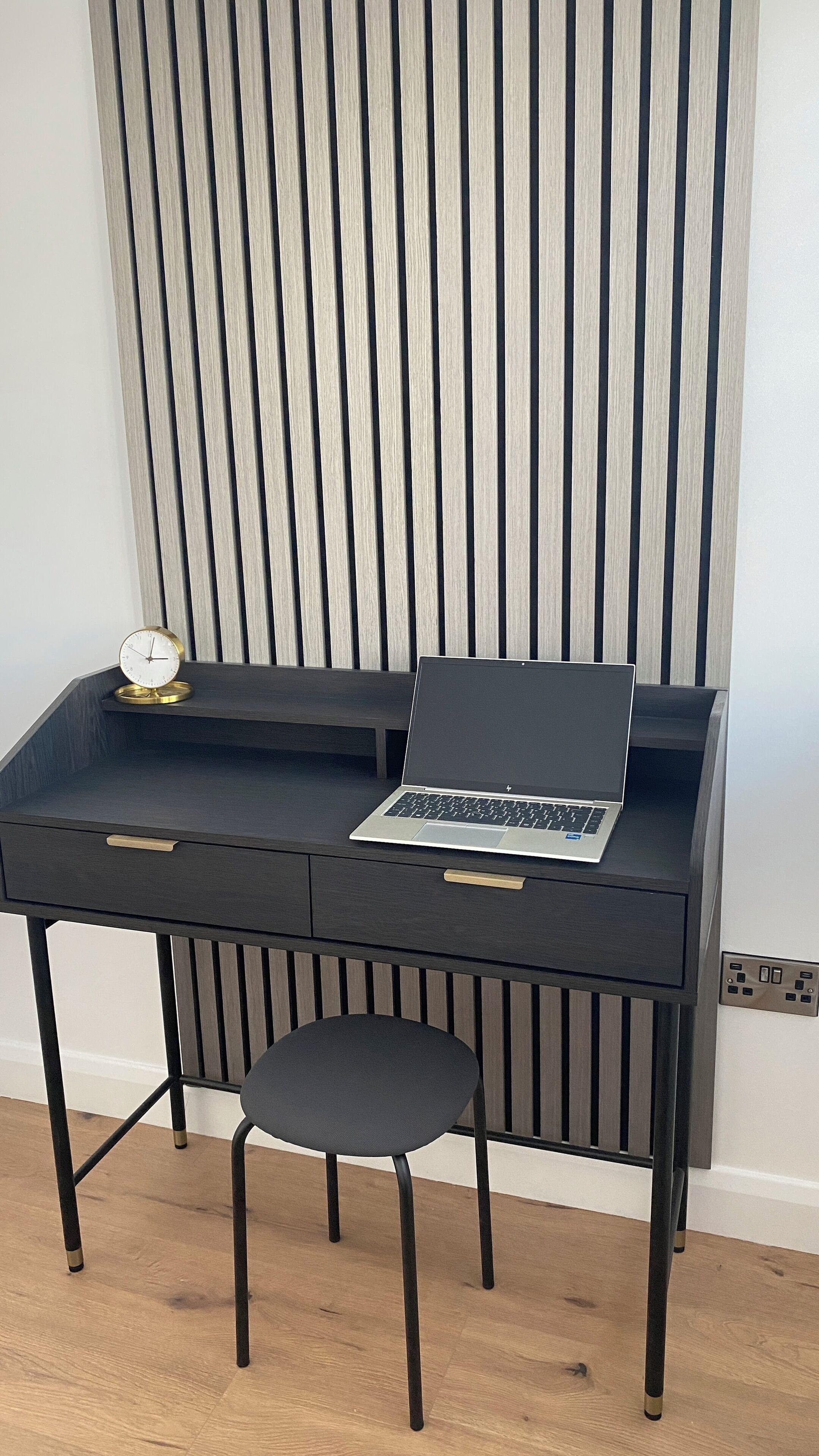 Un bureau en bois foncé, sur lequel se trouvent un ordinateur portable, un petit tabouret et une horloge.