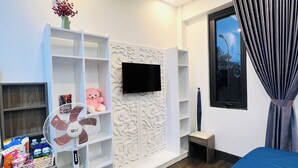 Une chambre à coucher moderne avec un lit, un téléviseur fixé au mur, un ventilateur sur pied et une petite table.