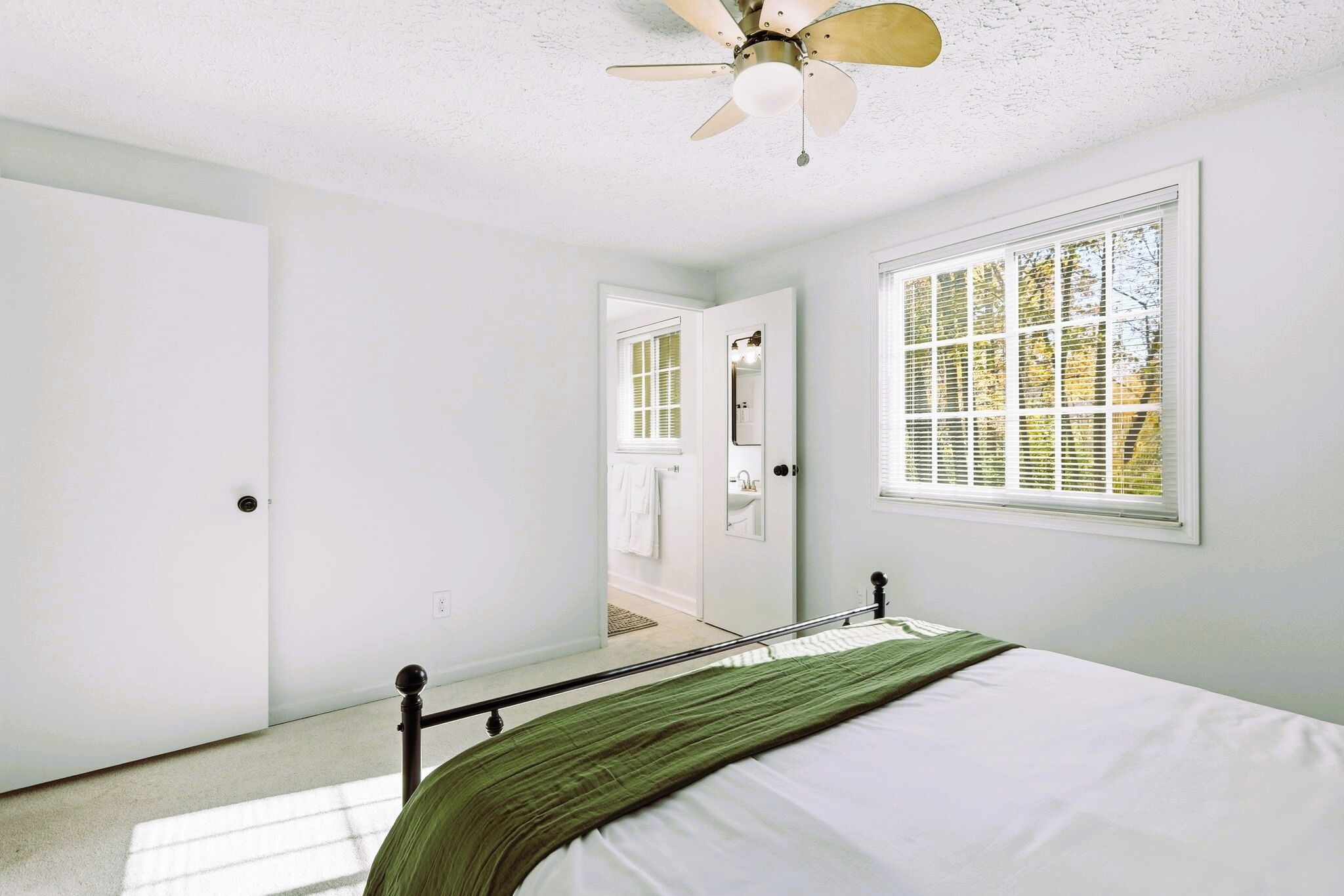 Une chambre à coucher comprenant un lit, un ventilateur de plafond, une fenêtre avec des stores et une porte donnant sur une salle de bain.