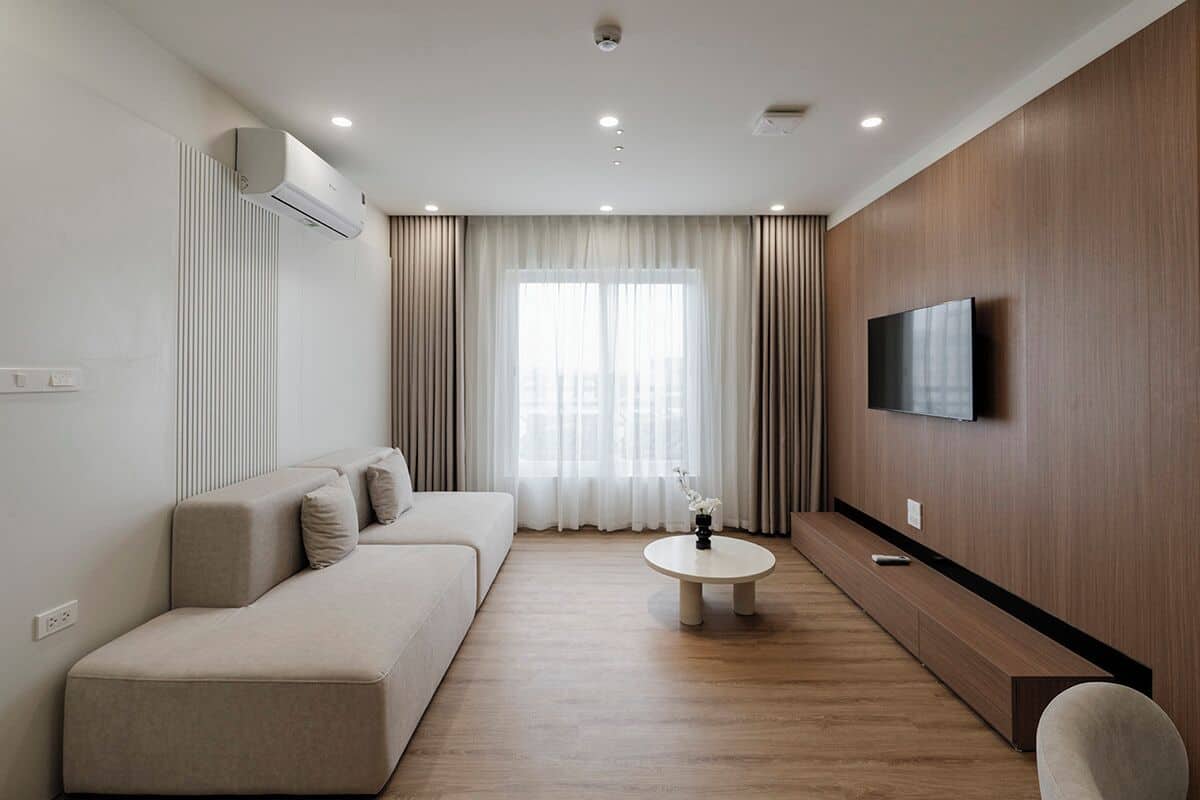 A modern living room with a sofa, a coffee table, a flat-screen TV mounted on the wall, and a large window with curtains.