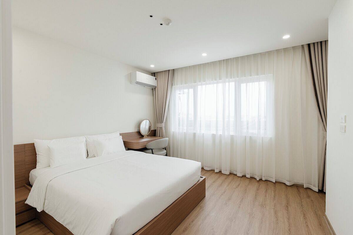 A modern bedroom with a wooden bed, a desk, and a large window with sheer curtains.