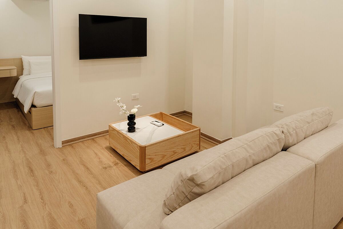 A modern hotel room with a wooden floor, a beige sofa, a small wooden side table with a vase of flowers, a flat-screen TV mounted on the wall, and a bed with white bedding.