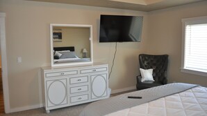 Un dormitorio con cama, cómoda, silla, televisor y ventana.