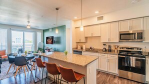 A modern kitchen with white cabinets, a stainless steel oven, and a granite countertop. There are two bar stools and a dining table with chairs.