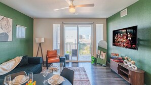A living room with a dining table set, a sofa, a TV mounted on the wall, and a ceiling fan.