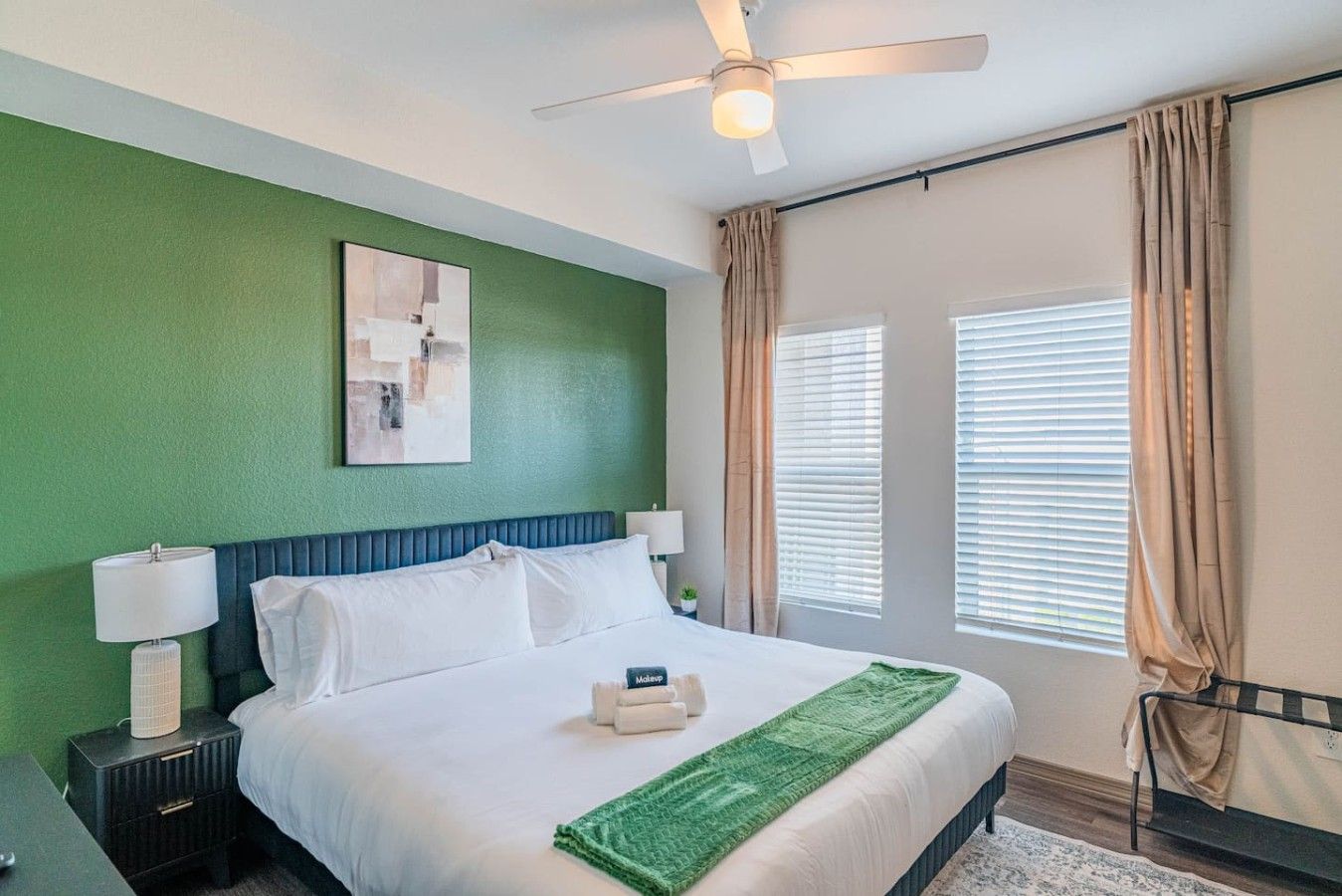 A hotel room with a bed, a ceiling fan, a nightstand, a lamp, a painting, and a window with curtains.
