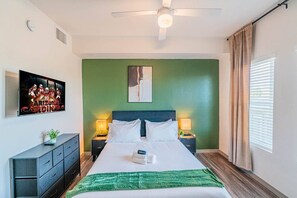 Ein Hotelzimmer mit einem Bett, zwei Nachttischen mit Lampen, einem Deckenventilator, einer grünen Wand, einem gerahmten Bild und einem Fenster mit Vorhängen.