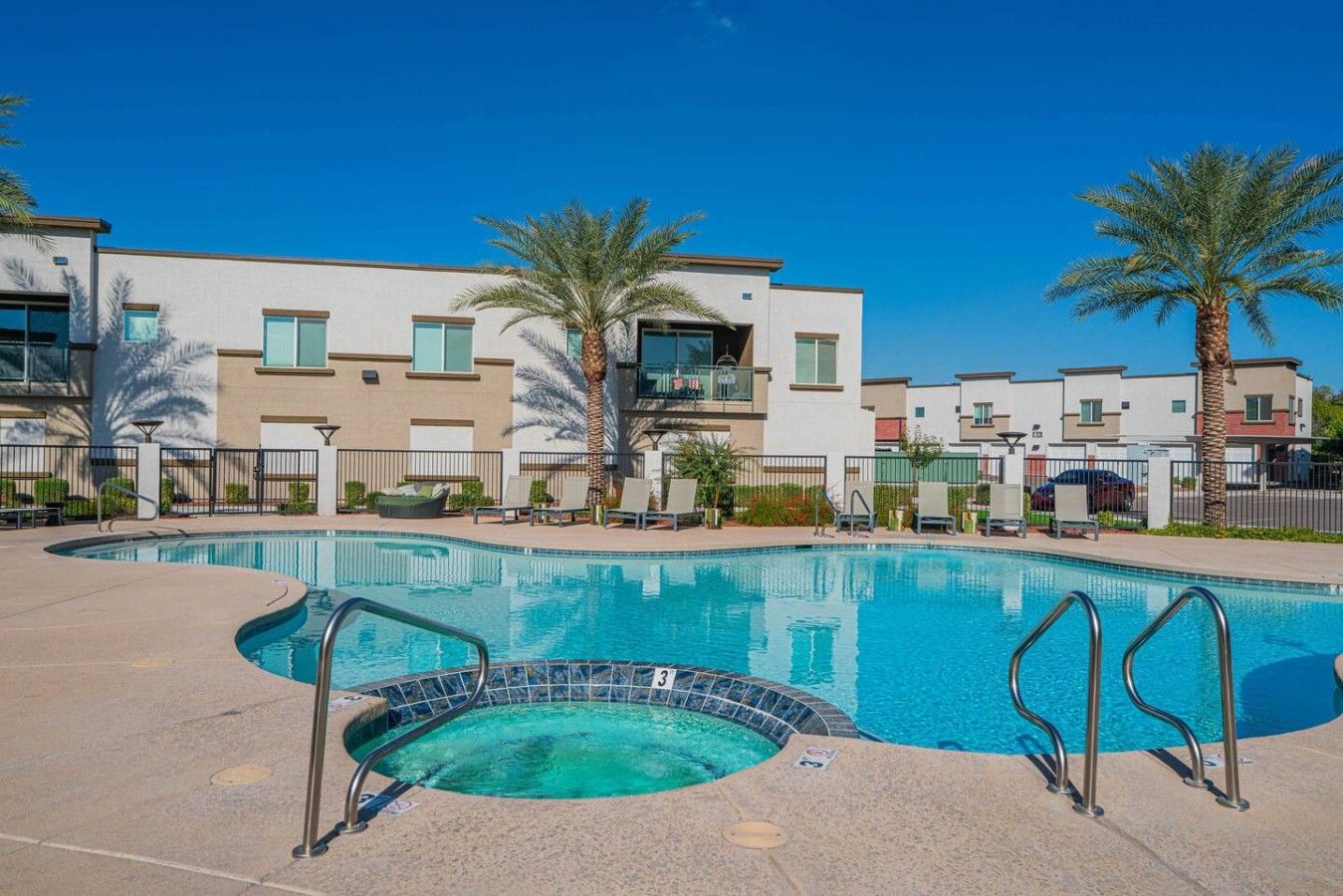 A swimming pool with a hot tub, surrounded by a fence, and adjacent to a row of modern apartment buildings.