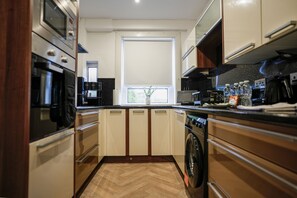 A compact kitchen with a built-in oven, microwave, and washing machine, a window with a view, and a herringbone-patterned floor.