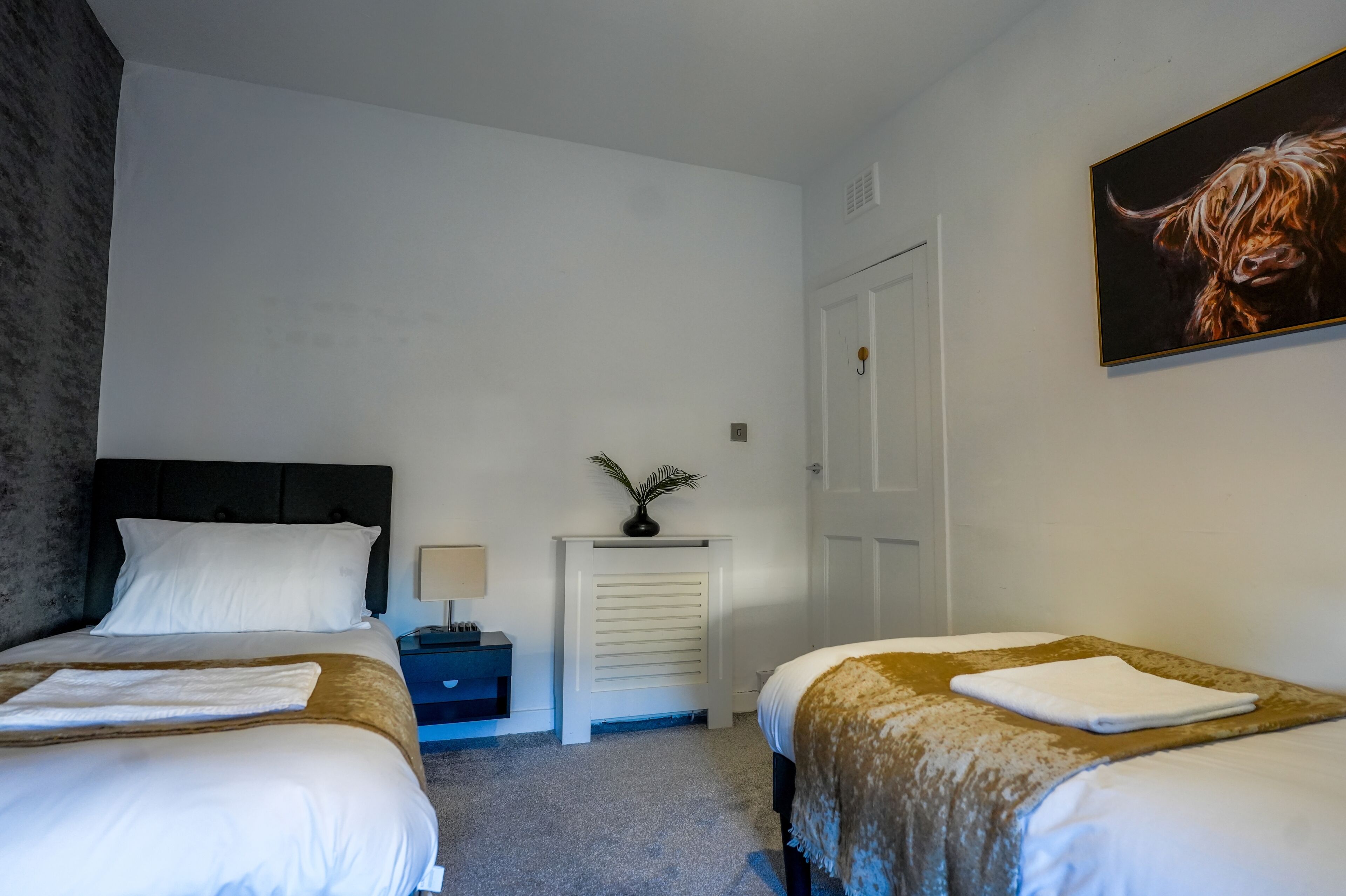 A hotel room with two beds, a nightstand, a radiator, a door, and a painting.