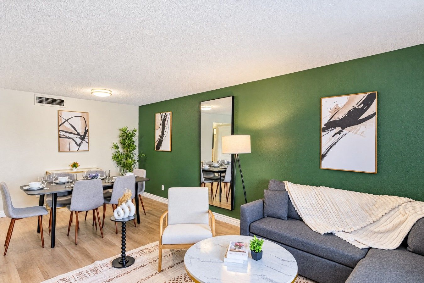 A modern living room with a green accent wall, a grey sofa, a dining area with a table and chairs, and artwork on the walls.