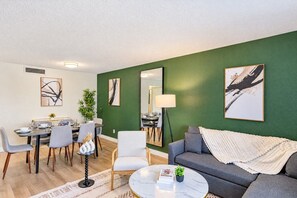 Una sala moderna con una pared verde de acento, un sofá gris, una zona de comedor con mesa y sillas, y obras de arte en las paredes.