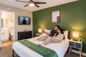 Une chambre à coucher avec un lit, des tables de chevet, un ventilateur de plafond, une télévision et un mur d’accent vert.