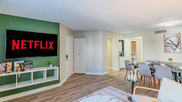 Een woonkamer met een flatscreen televisie, een boekenkast, een eethoek met een tafel en stoelen, en een gang die naar een keuken leidt.