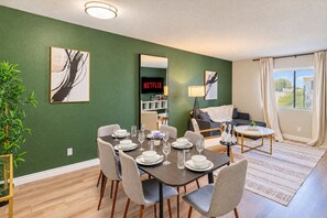 Un área de comedor con una mesa preparada para cuatro personas, un sofá, un televisor mostrando 'NETFLIX' y una ventana con cortinas.