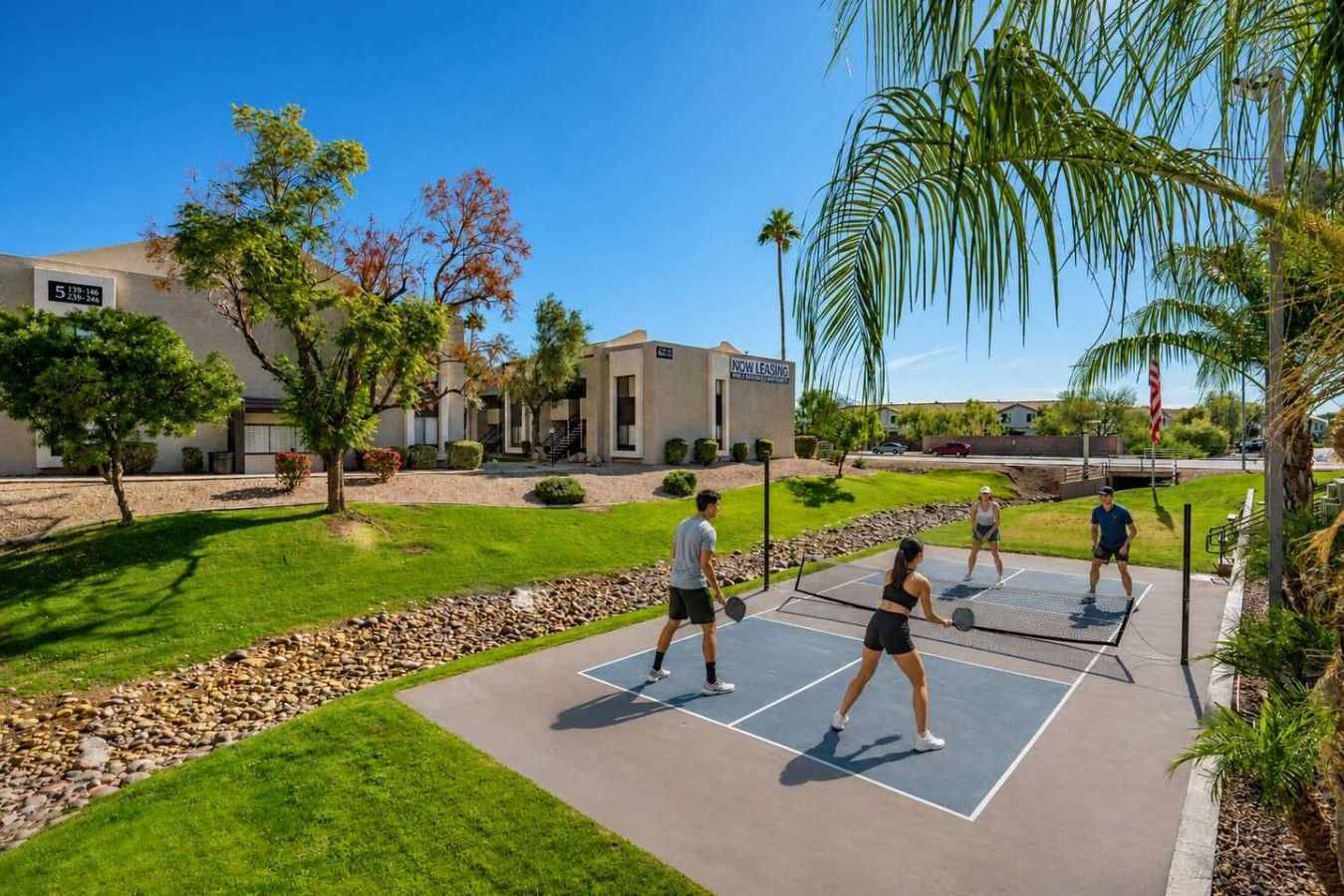 A tennis court with players, surrounded by a landscaped area with trees and a building labeled 'NOW LEASING'.