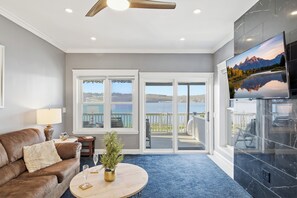 A living room with a brown leather sofa, a round coffee table, a small table with a lamp, and a potted plant. There is a large TV on the wall and a sliding glass door leading to a balcony with a view of the water. - Riverview Retreat: Guest Suite (Groton)