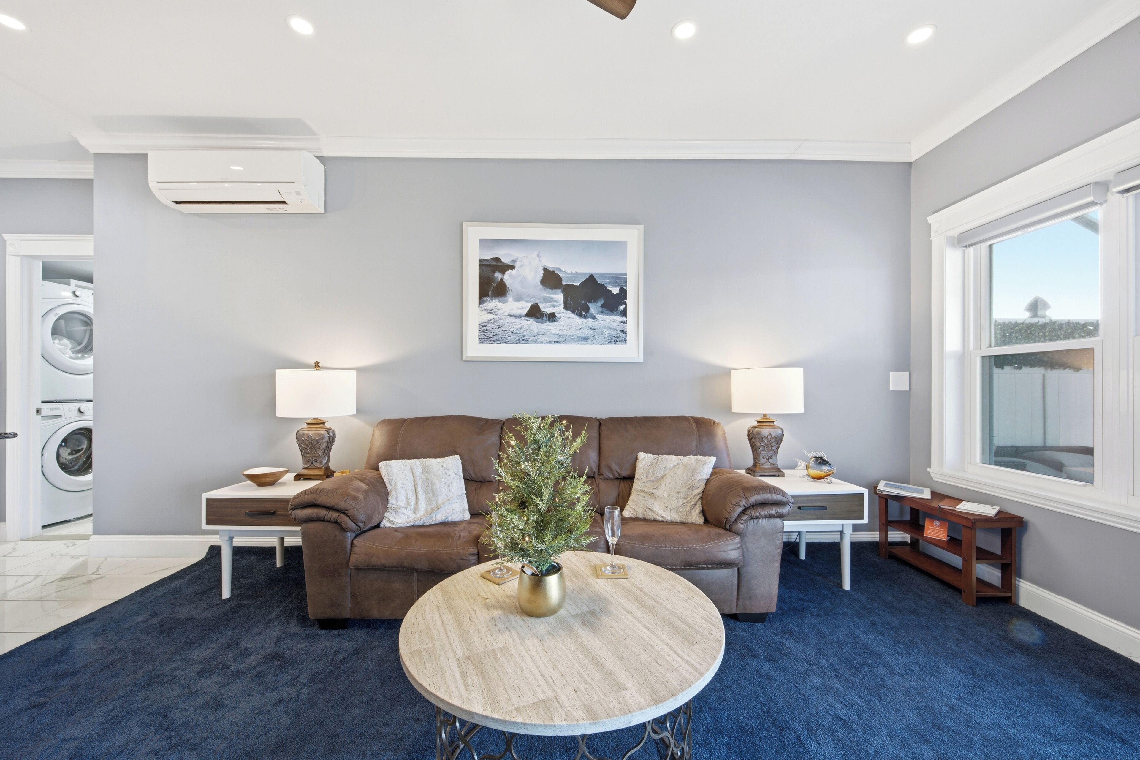 A living room with a brown leather sofa, two side tables with lamps, a round coffee table with a plant, and a framed picture on the wall.