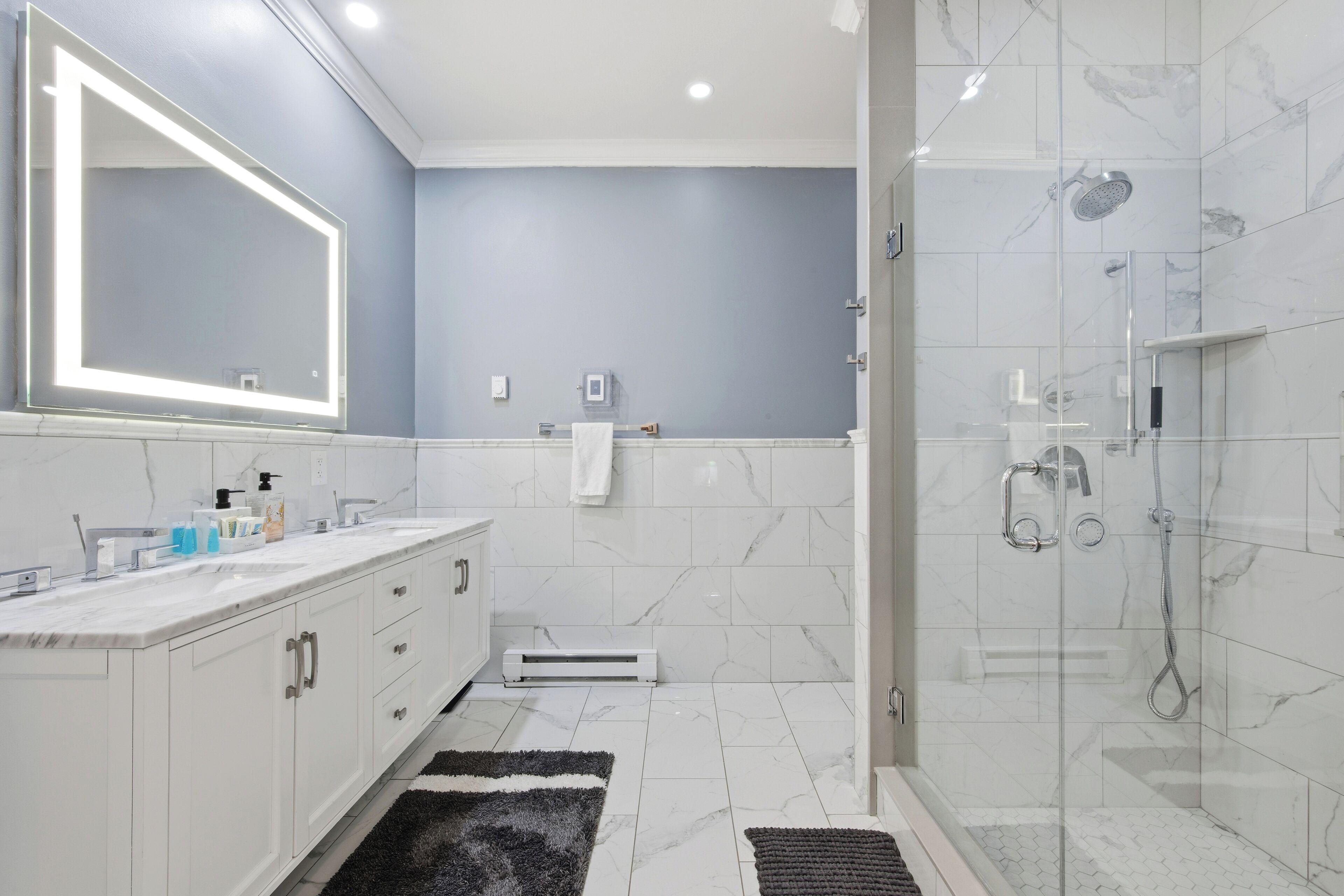 A modern bathroom with a large mirror, white marble tiles, a glass-enclosed shower, and a black rug.