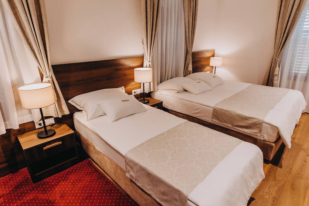 Een hotelkamer met twee eenpersoonsbedden, houten hoofdborden, nachtkastjes met lampen en een rood tapijt met dessin.
