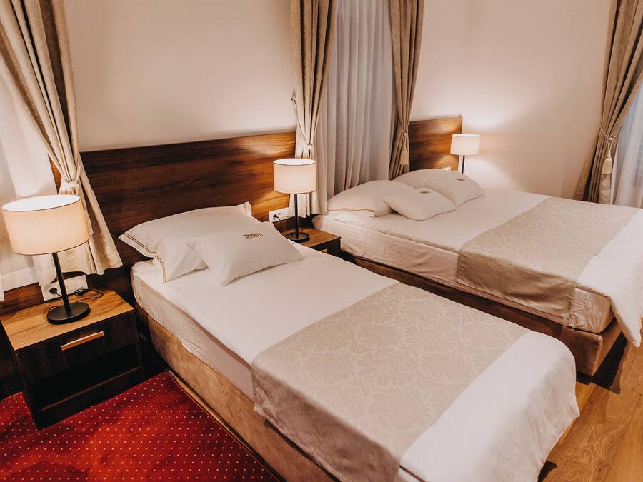 A hotel room with two single beds, wooden headboards, bedside tables with lamps, and a red patterned rug.