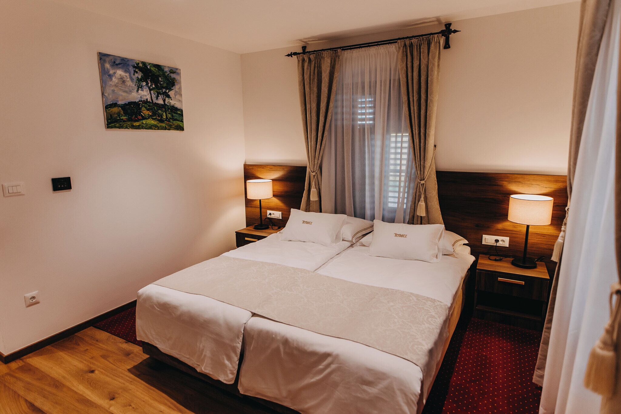 A double bed with two bedside tables, each with a lamp, a painting on the wall, and a window with curtains.