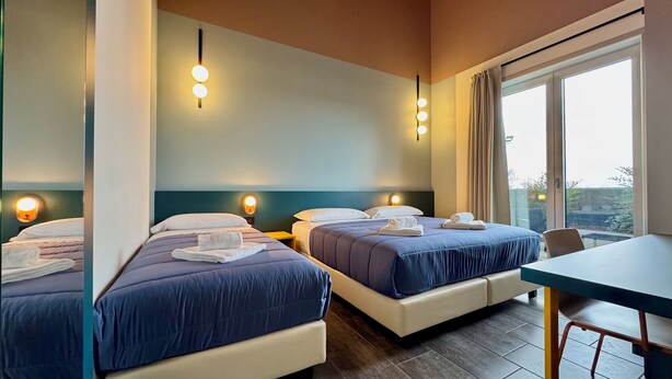 A hotel room with two beds, a table, and a large window. - HC Hotel Manerbio Loft (Manerbio)