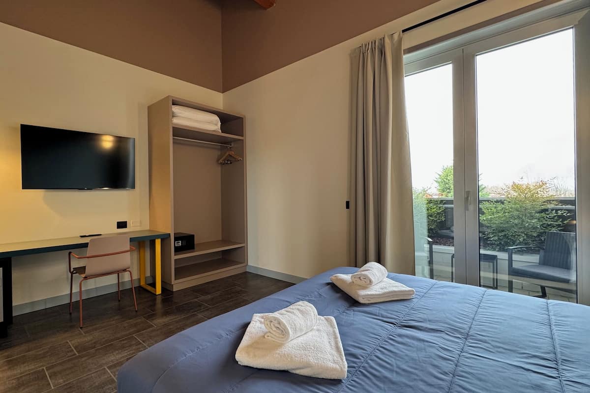 A hotel room with a bed, a desk, a chair, a TV, a wardrobe, and a balcony with a view.