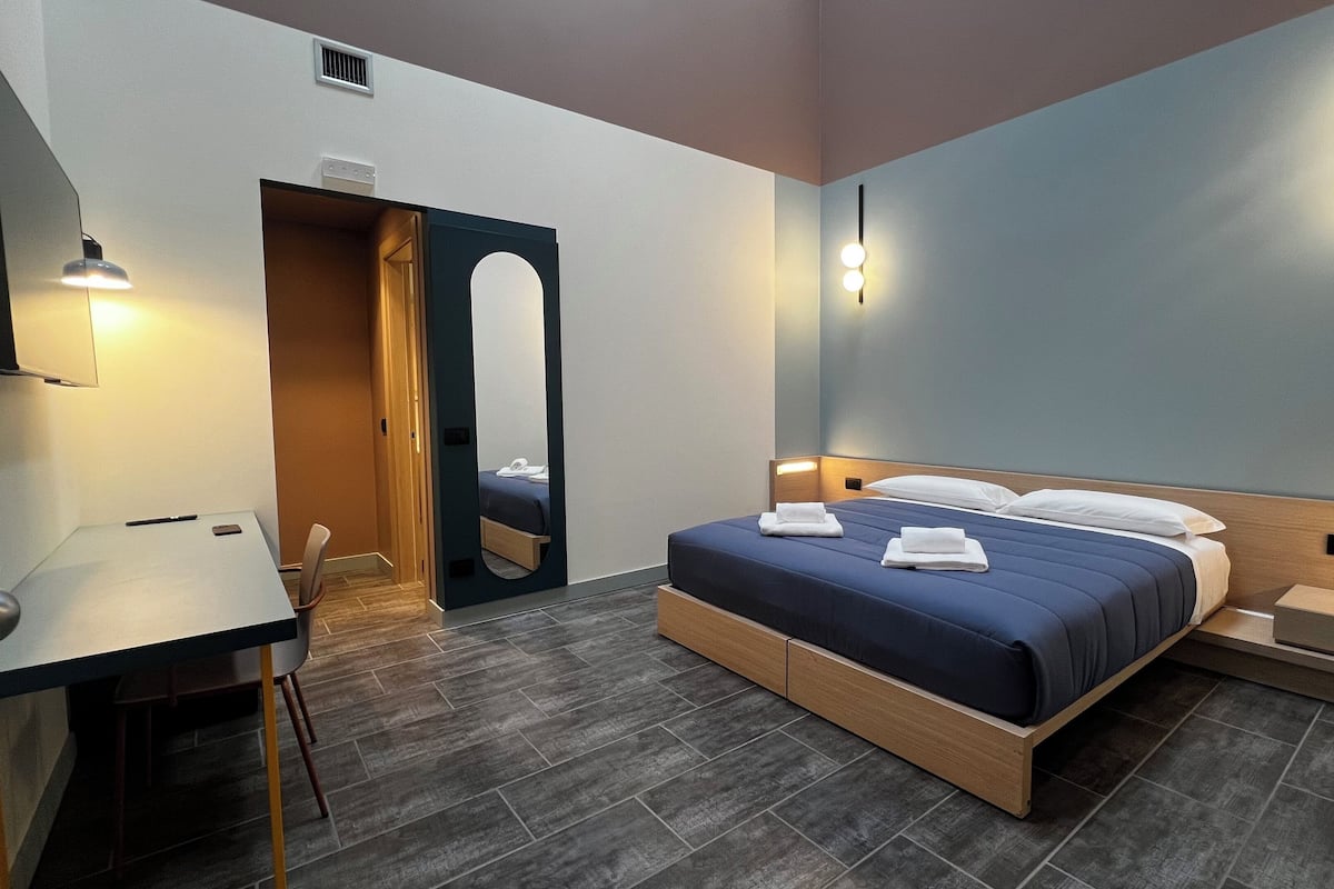 A modern hotel room with a bed, bedside tables, a desk, and a mirror.
