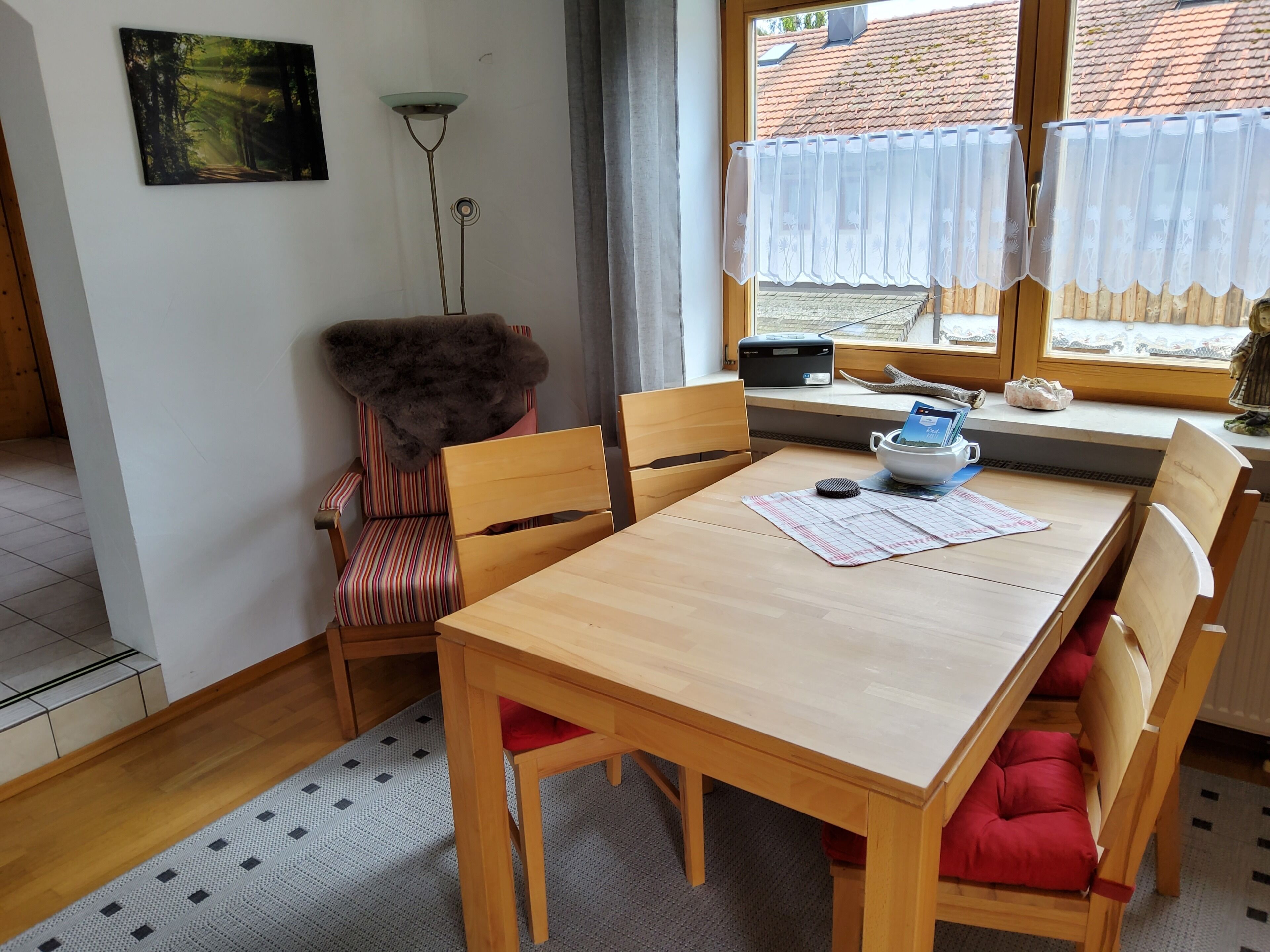 Un coin repas avec une table et des chaises en bois, un plaid en fourrure sur une chaise, une lampe sur pied et une fenêtre donnant sur un immeuble.