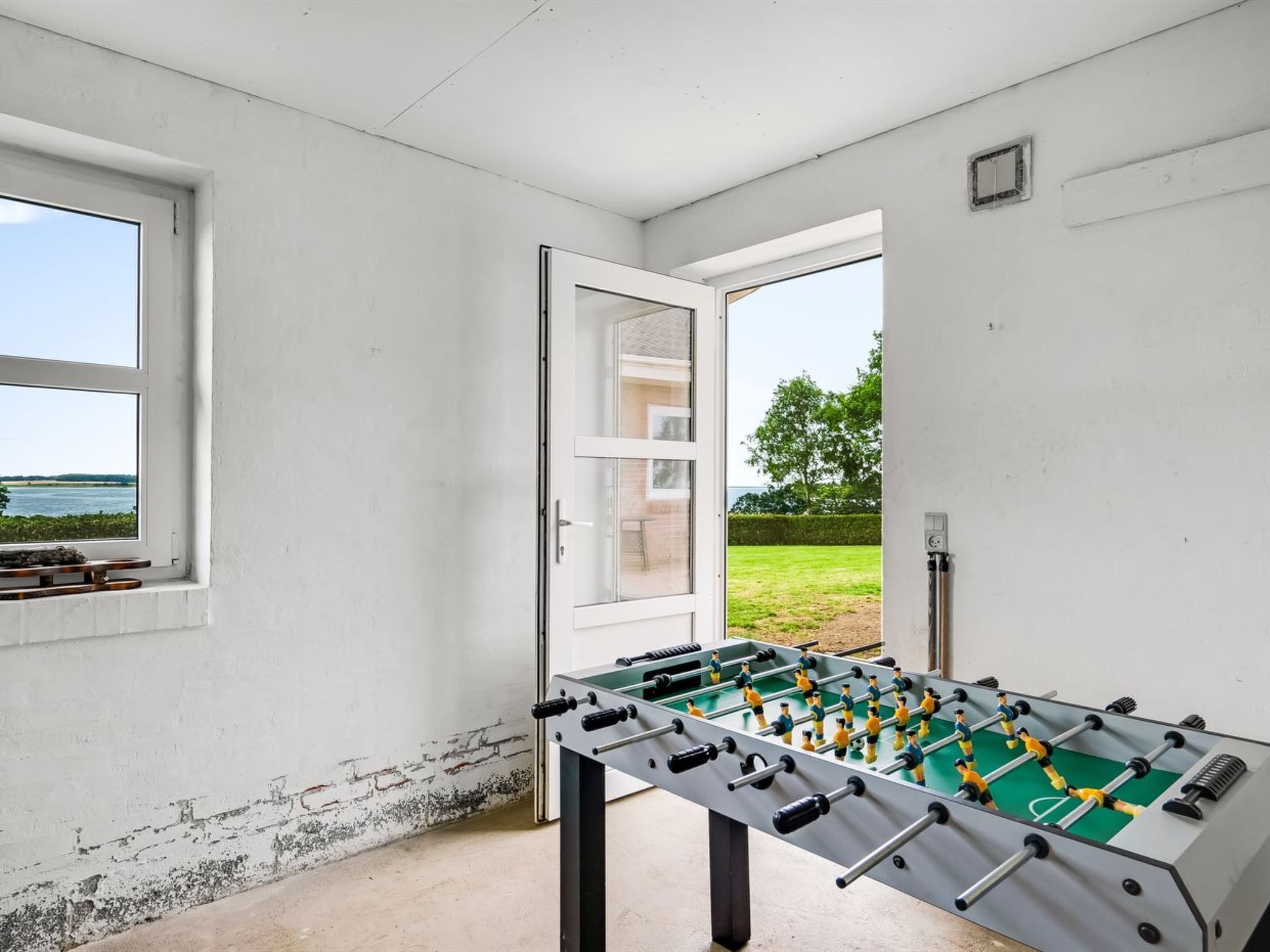 A room with a foosball table, a window with a view of a grassy area and trees, and a door leading outside.