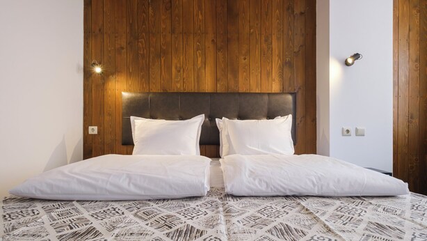 A double bed with a patterned bedspread, a wooden headboard, and two wall-mounted lights. - Hotel Razlog (Razlog)