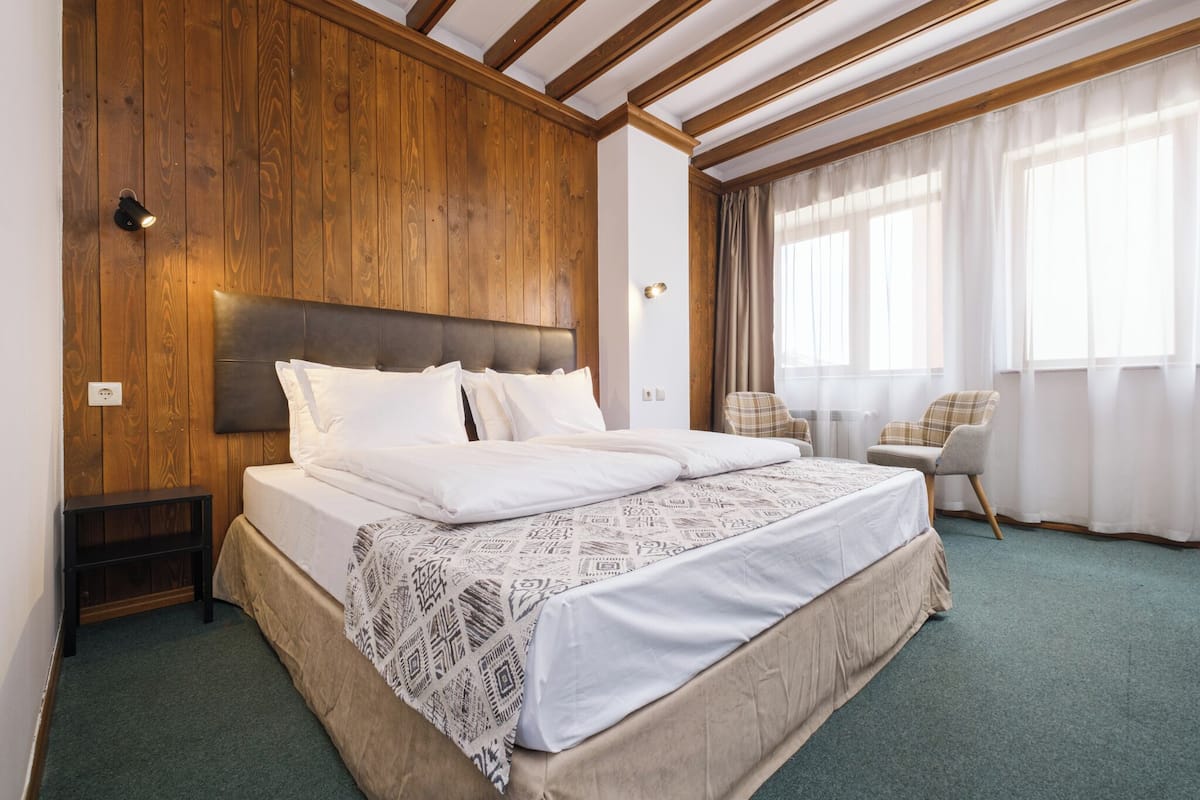 A bedroom with a wooden headboard, a bed with white linens, a small bedside table, a chair, and a window with curtains.