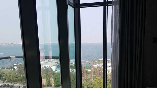 A room with a large glass window offering a view of a body of water and a distant cityscape. - Oceanview apartment with FREE parking in Hoora Manama (hoora manama)