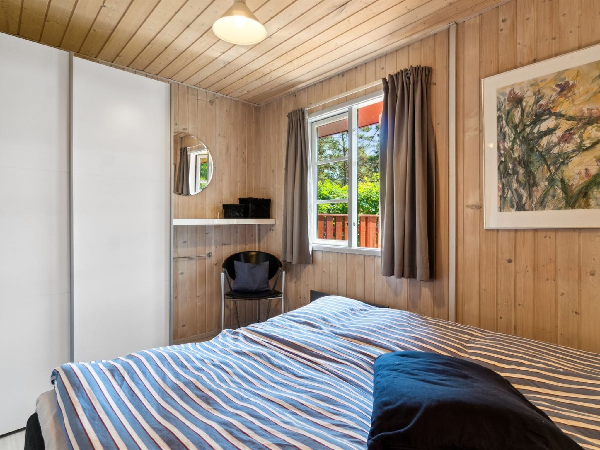 Ein Schlafzimmer mit einem Bett, einem Sessel, einem Regal mit Körben, einem Fenster mit Vorhängen und einem Bild an der Wand.