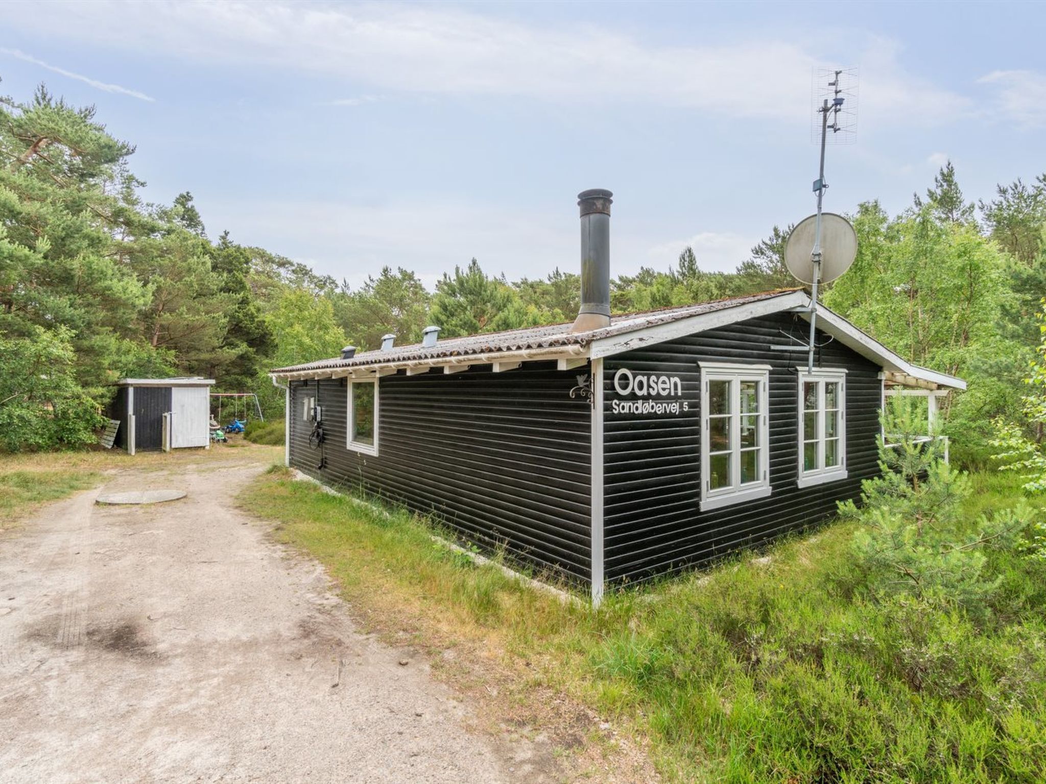 A small, single-story cabin with a chimney and satellite dish, surrounded by trees and a gravel path.