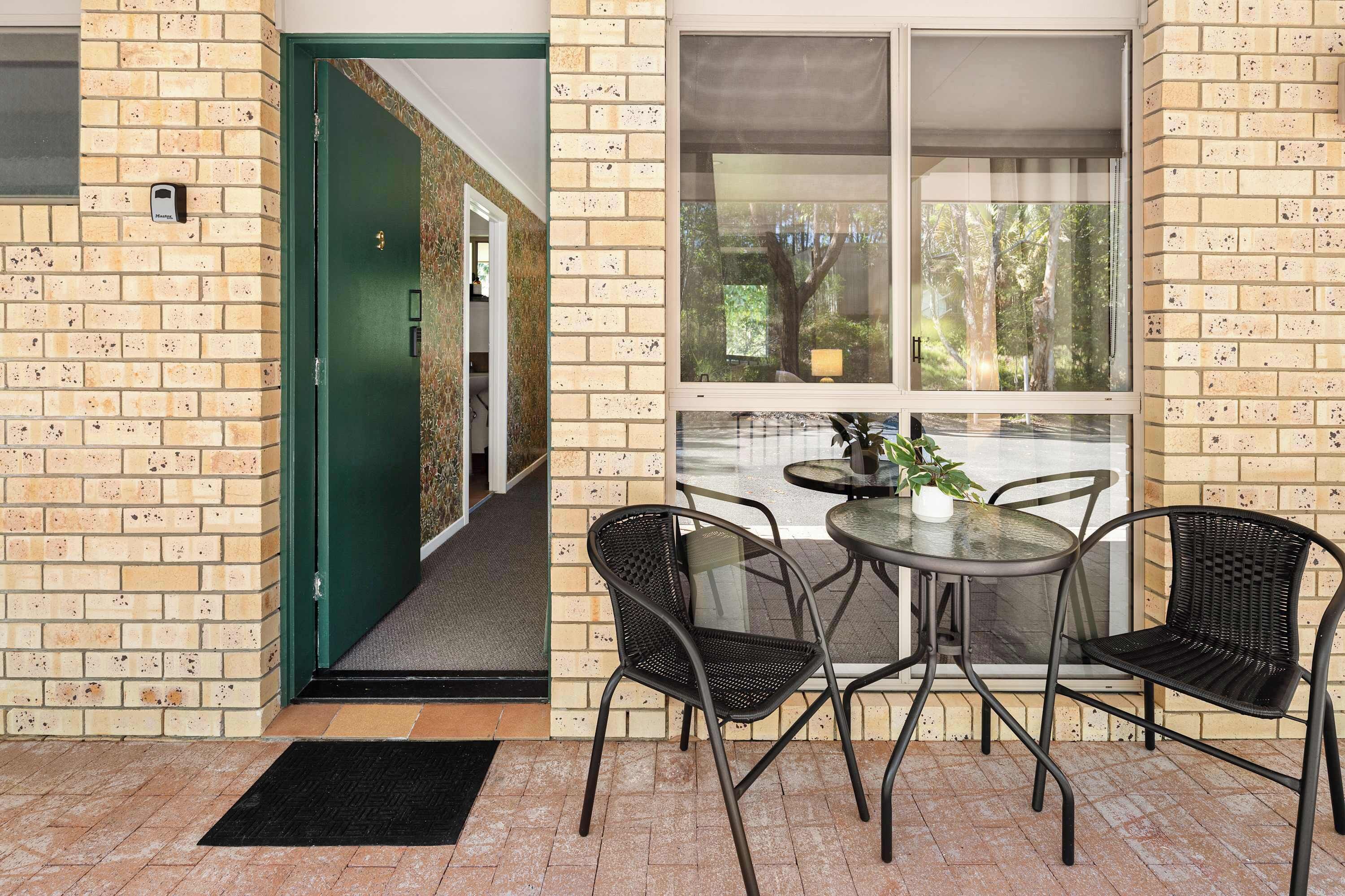 A patio area with a table and chairs, a green door, and a window with a view of trees.