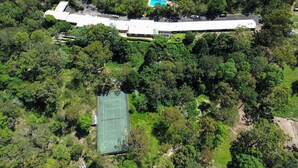 Vue aérienne d’un bâtiment moderne doté d’un long toit incurvé, entouré d’une végétation luxuriante et d’un court de tennis.