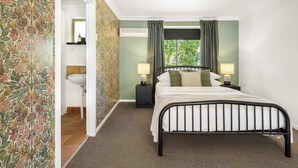 Une chambre à coucher comprenant un lit, des tables de chevet, une lampe, une fenêtre avec des rideaux, et une salle de bain avec un lavabo et un miroir.