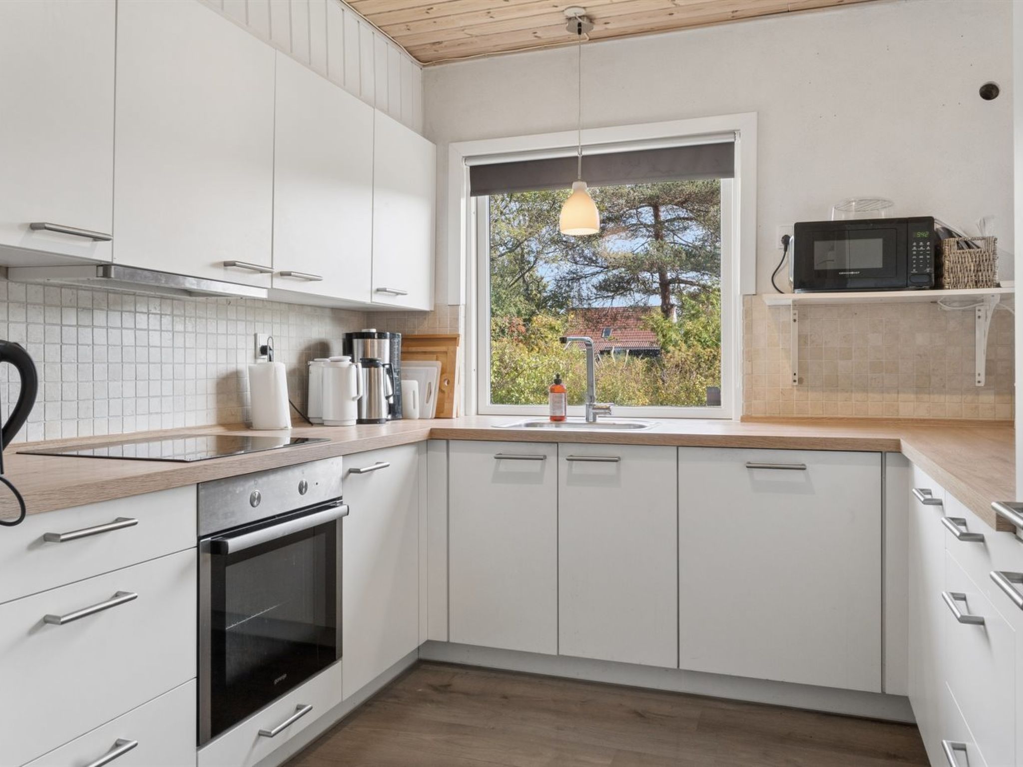 A modern kitchen with white cabinets, a built-in oven, a microwave, and a window with a view of trees.
