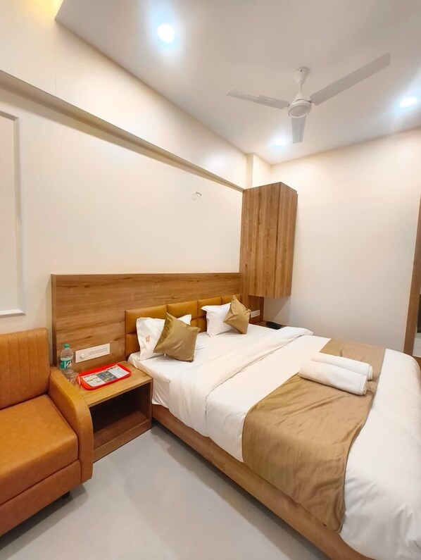 A hotel room with a wooden headboard, a bed with white and beige bedding, a nightstand with a red tray, and a ceiling fan. - Hotel Vistara (Ujjain)